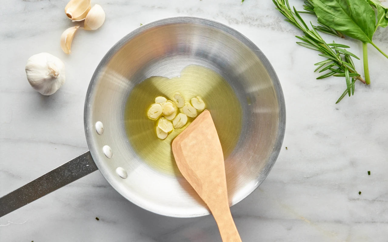 A stainless steel saucepan with a wooden spoon on a light marble surface, containing oil and garlic cloves, with garlic, basil, and rosemary arranged around it.