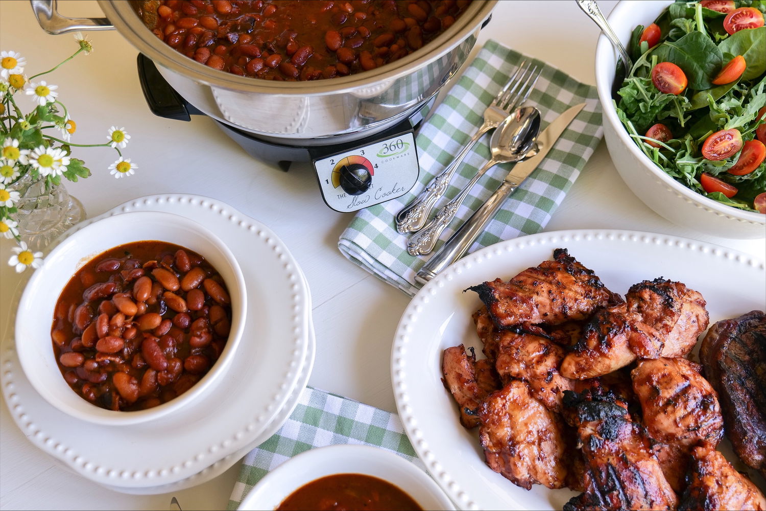 Table with food including grilled chicken, beans, salad, and a 360 Cookware slow cooker.