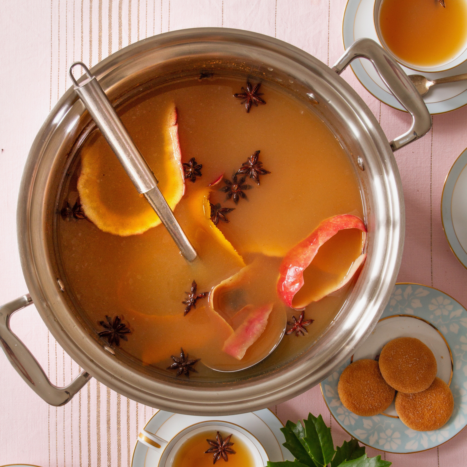 A 12-quart stainless steel stock pot filled with soup, garnished with apple peels and star anise, on a table with plates and cookies.