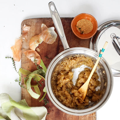 Food cooking in a 3 quart stainless steel pot with lid off to the side next to onion and apple peels
