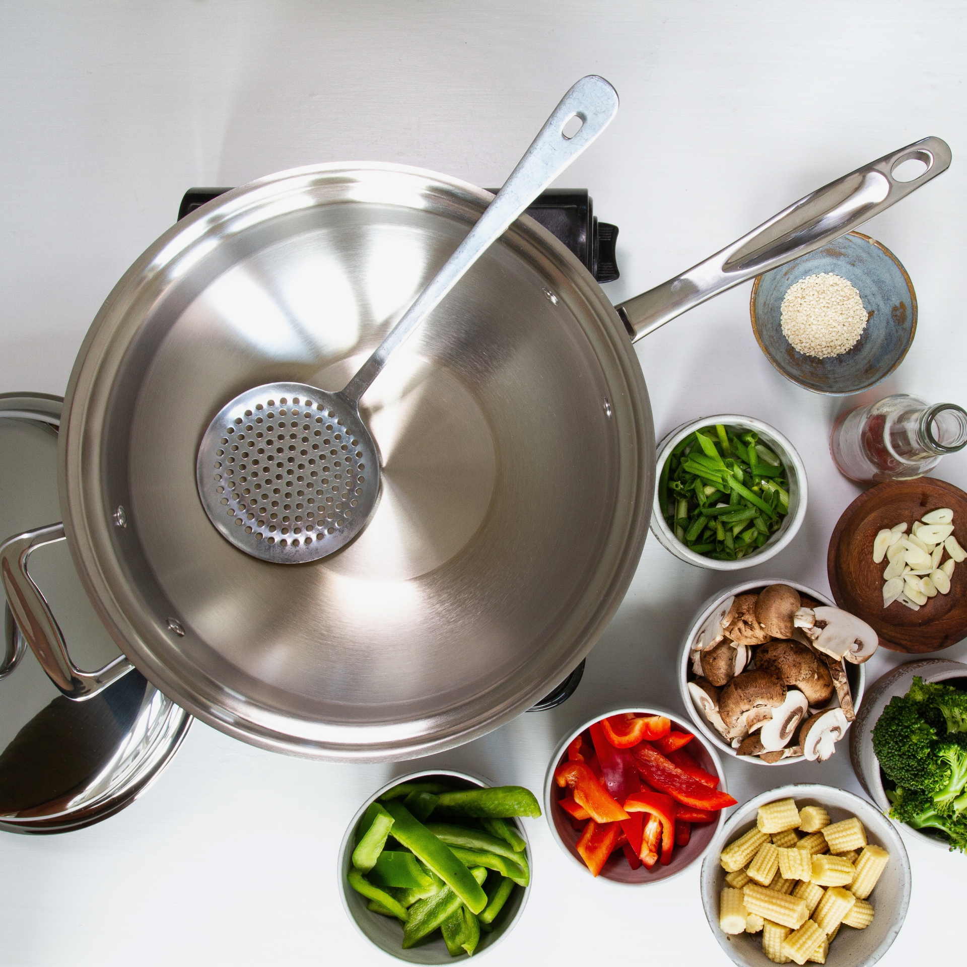 Stainless-steel wok with vegetables and a strainer on a white surface.