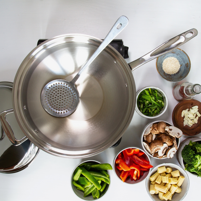 Stainless-steel wok with vegetables and a strainer on a white surface.