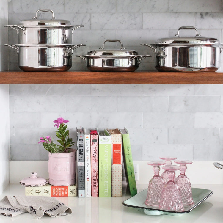 Stainless steel cookware on a shelf above cooking books and glassware