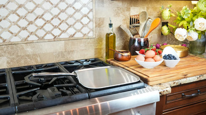 Angled view of 11-inch stainless steel square griddle with flat cooking surface and long riveted handle on a gas stove