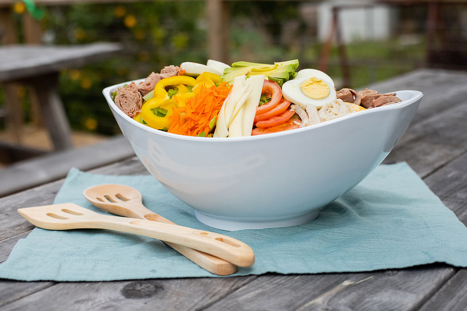 White salad bowl filled with chef's salad, including vegetables and noodles, on a table with wooden utensils.
