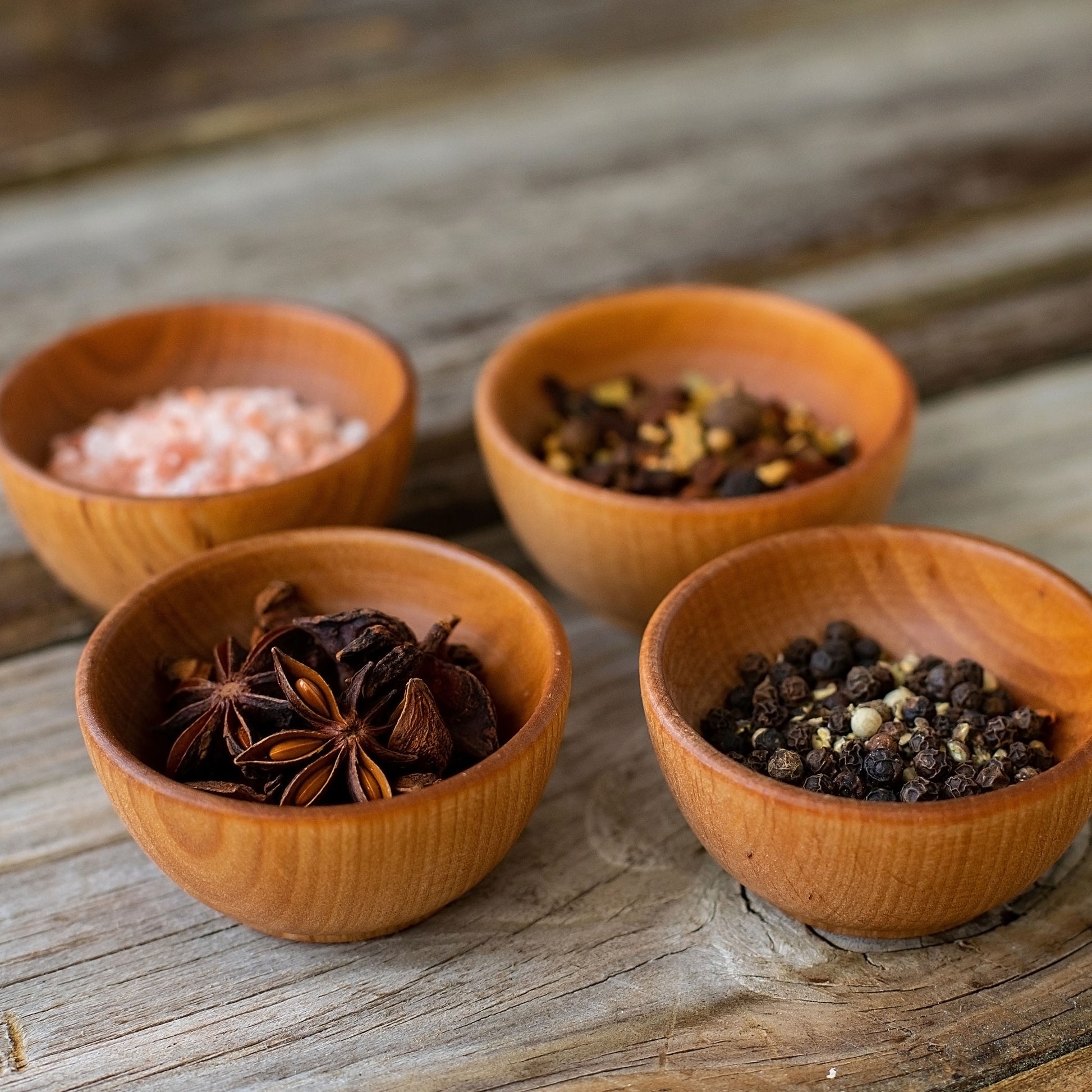 Four wooden bowls with different spices: pink salt, mixed spices, star anise, and black peppercorns.