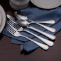 Polished stainless flatware set (forks and spoons) arranged on a folded blue cloth napkin on a wooden table.