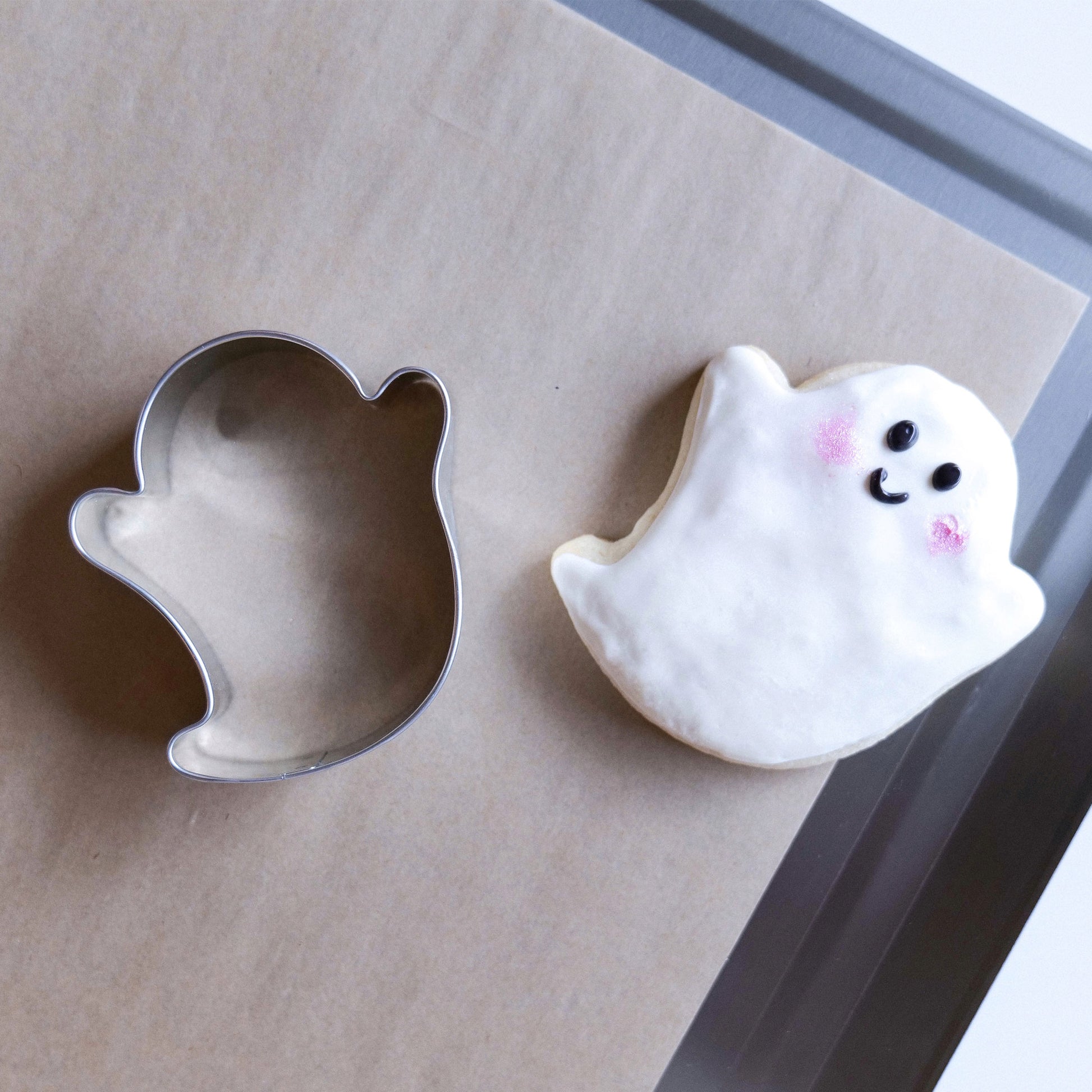 Ghost-shaped cookie cutter next to a decorated ghost cookie on parchment paper.