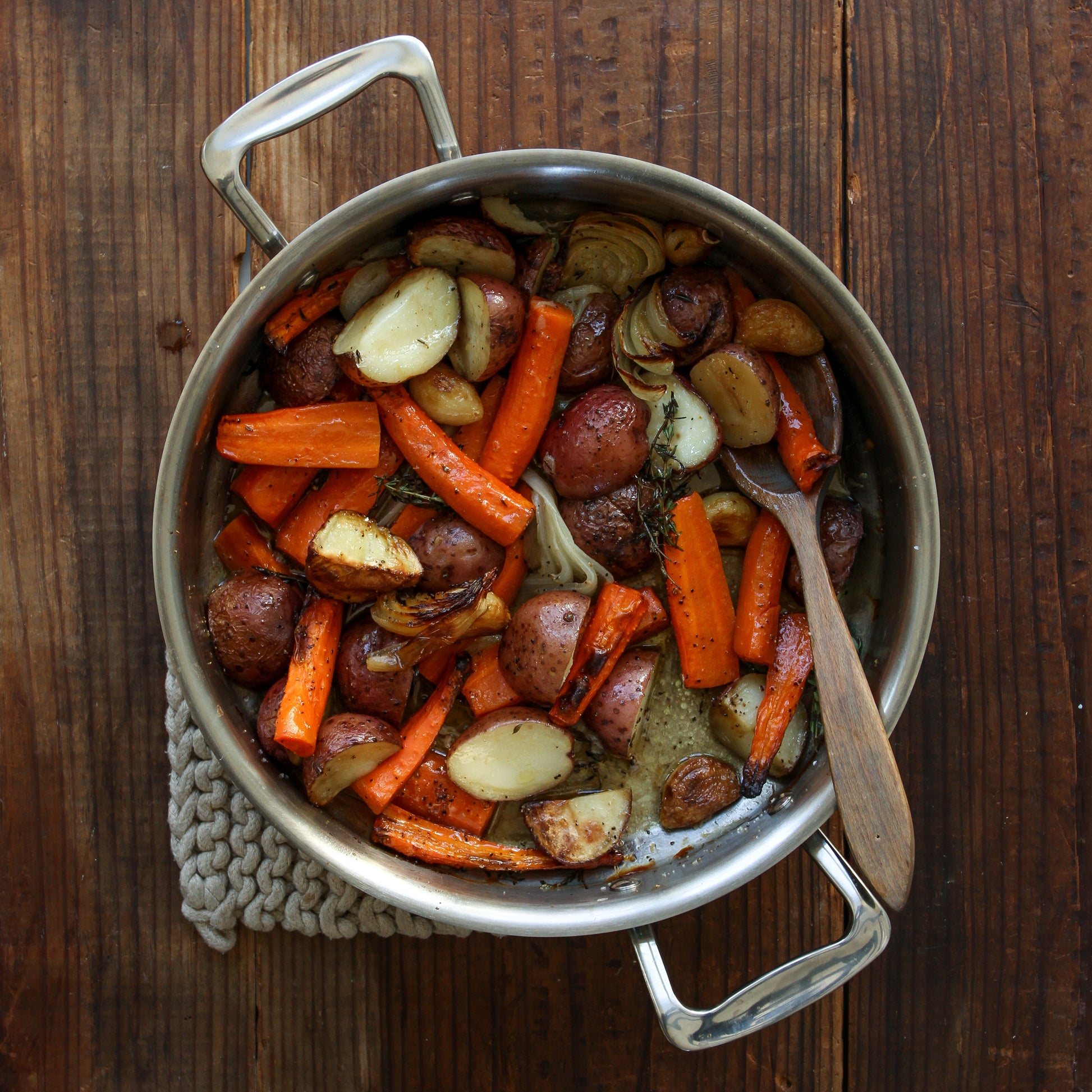 A sauté pan filled with roasted carrots, potatoes, and onions on a wooden surface.
