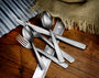 Stacked stainless-steel flatware (fork, spoons, knives) on a wooden table with a burlap cloth.
