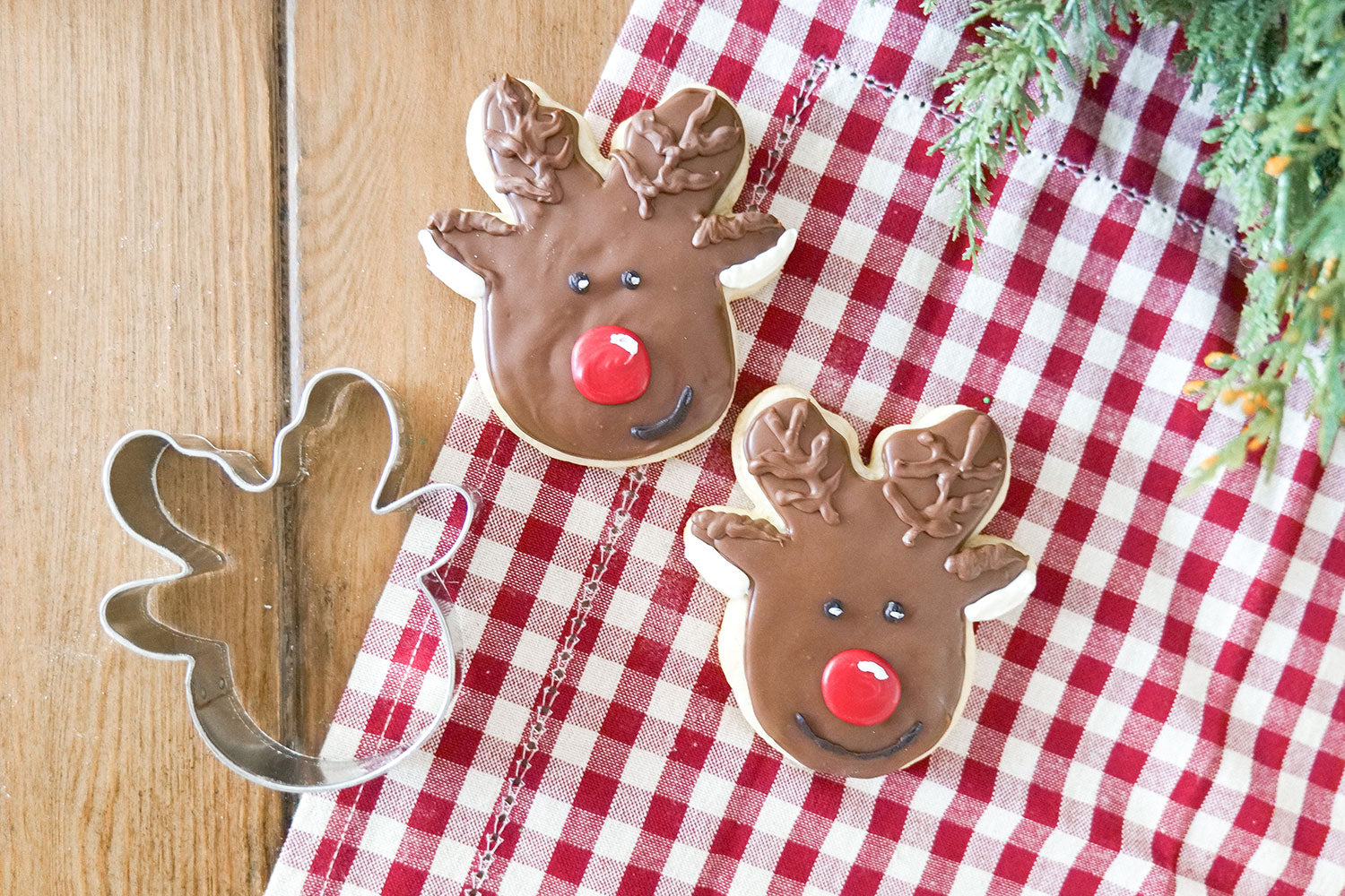 Two decorated reindeer cookies with a reindeer cookie cutter on a checkered cloth.