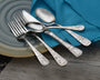 Stacked ornate silver fork and spoons with engraved handles on a gray plate beside a teal cloth napkin on wood table.