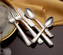 Angled view of Prestige flatware showing feather-border handles and polished 18-10 stainless on dark table with gold napkin