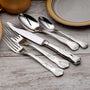 Angled view of Sheffield- 65 Piece Set highlighting ornate 18-10 stainless handles on weathered wood beside a plate