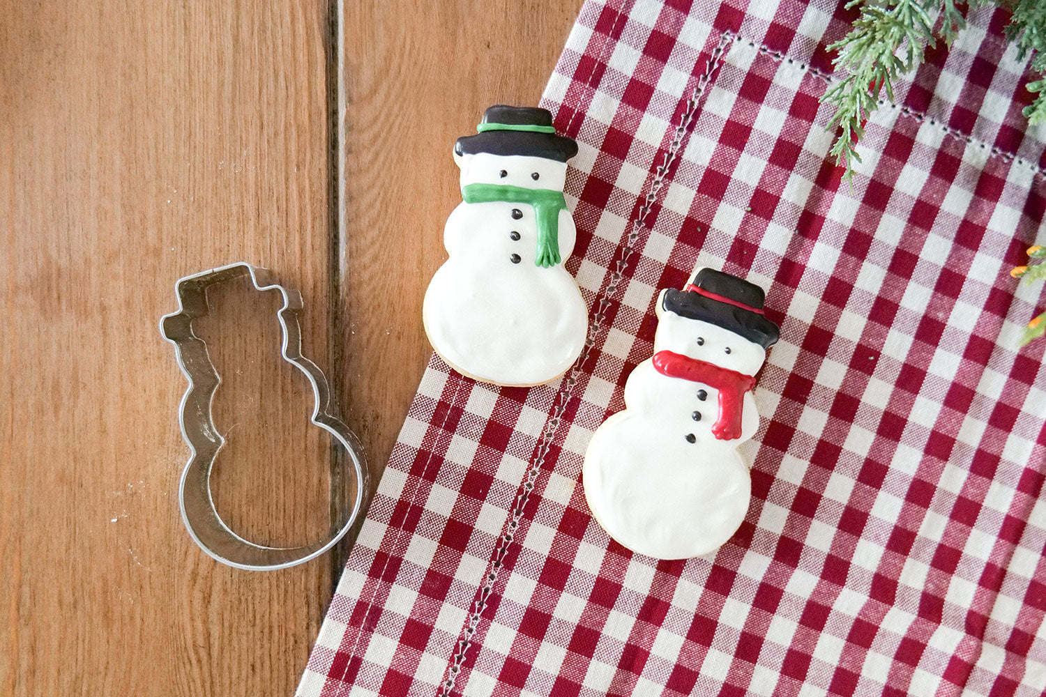 Two decorated snowman cookies with a snowman cookie cutter on a checkered tablecloth.